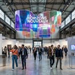 Bustling art fair in an industrial hall. A large, iridescent 'Horizons Nouveaux' installation hangs above diverse visitors viewing modern art.
