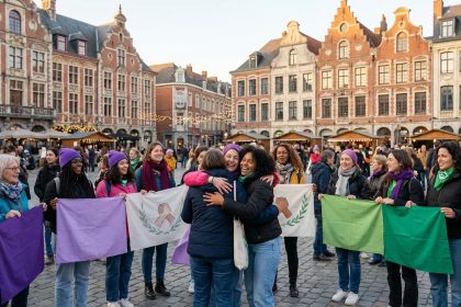 Groupe de femmes diverses souriantes, tenant des drapeaux violets, verts et blancs avec un symbole de mains serrées, dans une place de ville animée.