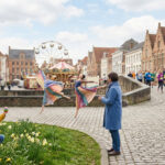 Scène de rue animée d'une ville européenne au printemps : ballerines, coureurs, enfant et grande roue, sous un ciel nuageux.
