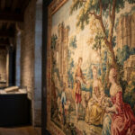 Tapisserie du 18e siècle en gros plan, avec scène pastorale, personnages en costumes, arbres et ruines. Au fond, musée avec visiteurs.