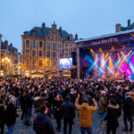Une foule dense danse devant une grande scène de concert illuminée sur la Grand-Place de Lille à la tombée de la nuit.