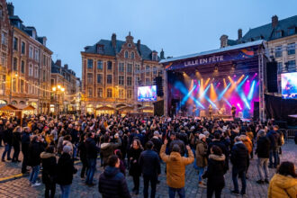 Une foule dense danse devant une grande scène de concert illuminée sur la Grand-Place de Lille à la tombée de la nuit.