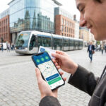 Un jeune homme tient un smartphone affichant l'application Ilevia devant un tramway à Lille.